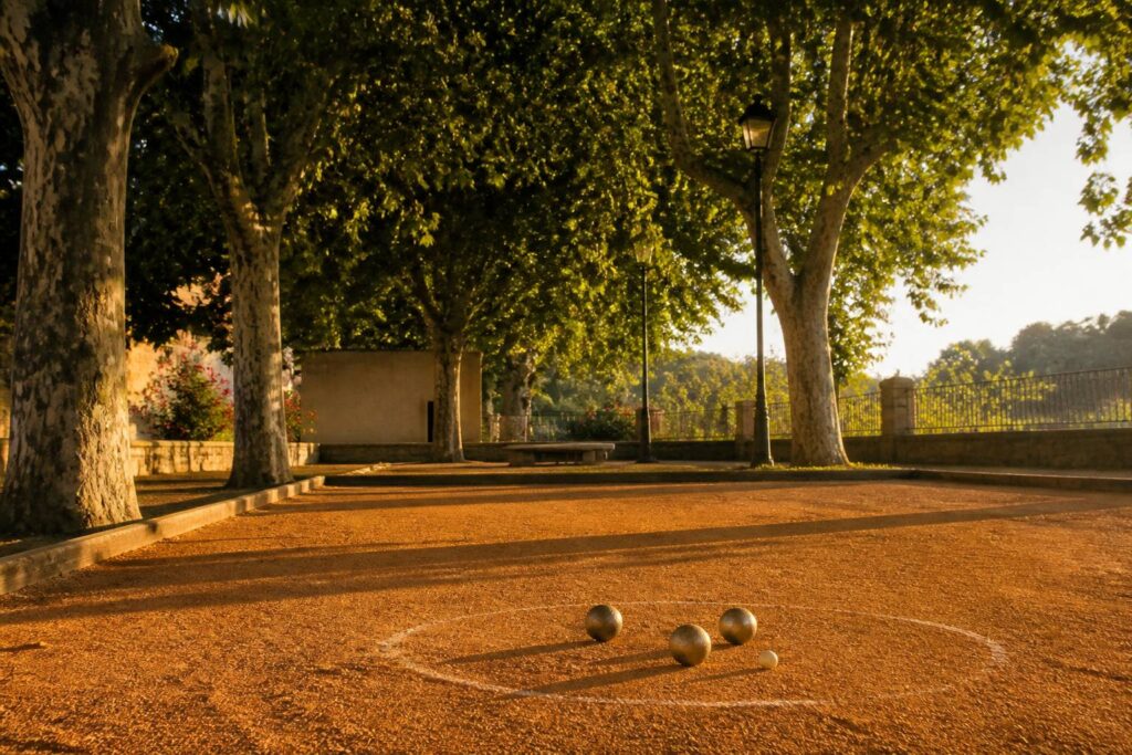 Terrain de pétanque en terre battue avec boules, cochonnet et platanes en fin d'après-midi